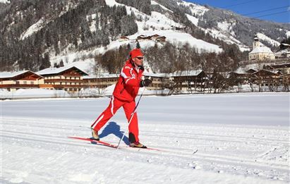 Eine Person in roter Ski-Ausrüstung fährt Langlauf auf einer verschneiten Strecke. Im Hintergrund sind schneebedeckte Berge und traditionelle chalets zu sehen.