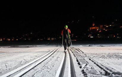 Eine Person mit Skistöcken fährt bei Nacht auf einer schneebedeckten Strecke. Im Hintergrund sind beleuchtete Gebäude zu sehen.