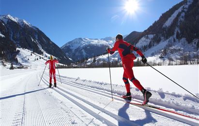 Zwei Skilangläufer in roten Anzügen bewegen sich auf einer verschneiten Strecke. Im Hintergrund sind schneebedeckte Berge und ein klarer blauer Himmel zu sehen.