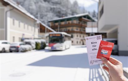Ein Blick auf eine verschneite Straße mit einem Bus und mehreren Gebäuden im Hintergrund. In der Hand hält jemand zwei Informationskarten.