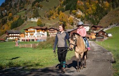 Eine Frau führt ein Pony, während ein Kind darauf sitzt. Im Hintergrund sieht man grüne Wiesen und traditionelle Häuser in einer malerischen Berglandschaft.