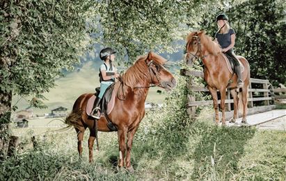 Zwei Personen reiten auf braunen Pferden in einer grünen Landschaft. Im Hintergrund sind Bäume und eine Wiese zu sehen.