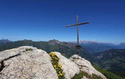 Ein wunderschöner Aussichtspunkt mit einem Holzkreuz auf einem felsigen Gipfel. Im Hintergrund erstrecken sich grüne Berge unter einem klaren blauen Himmel.