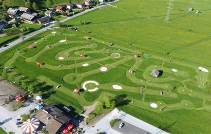 Eine große, grüne Wiese mit einer aufregenden Kartbahn, die in geschwungenen Linien angelegt ist. Im Hintergrund sind Häuser und ein kleiner Vergnügungsbereich zu sehen.
