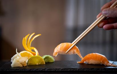 Ein kunstvoll angerichtetes Sushi-Plateau mit Lachs, Ingwer und Limette. Ein Hand mit Essstäbchen hebt ein Stück Sushi.