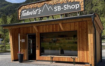 Ein Holzgeschäft mit dem Namen "Talwirt's SB-Shop" steht in einer grünen Landschaft. Es hat große Fenster und eine einladende Fassade.