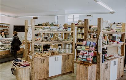 Ein gemütlicher Laden mit Holzmöbeln, der eine Vielzahl von Lebensmitteln und Produkten anbietet. Kunden shoppen zwischen den Regalen.