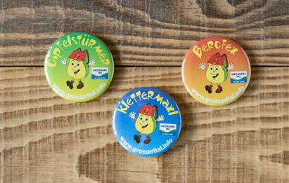 Drei bunte Anstecknadeln liegen auf einem Holztisch. Jede Nadel zeigt ein fröhliches Gesicht und einen unterschiedlichen Schriftzug.