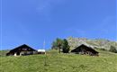 Zwei traditionelle Hütten auf einer grünen Wiese mit einem klaren blauen Himmel im Hintergrund. Die Umgebung ist von sanften Hügeln und Bergen geprägt.