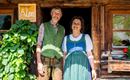Ein älteres Paar steht vor einer rustikalen Holzhütte. Beide tragen traditionelle Kleidung und lächeln freundlich in die Kamera.