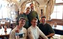 Eine Gruppe von fünf Personen lächelt in einem gemütlichen Restaurant. Die Atmosphäre ist freundlich und einladend, mit holzernen Tischen und Fenstern, die viel Licht hereinlassen.