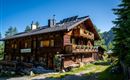 A charming wooden house in Alpine style with balconies and a sunny garden. Surrounded by green trees and a clear blue sky.