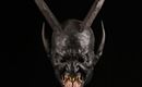 A mask-like face with long horns and a protruding tongue mouth. The depiction appears dark and eerie against a black background.