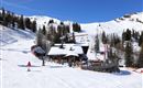An idyllic ski chalet in the middle of the snow with many skiers. In the background, there are snow-covered mountains and a blue sky.