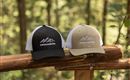 Two caps are standing on a wooden railing in the forest. One cap is black and the other is khaki, both featuring a mountain motif and the inscription "GREAT".