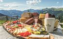 A wooden board with a selection of cheese varieties and sausage, surrounded by mountains and a blue sky. In the foreground, there is a cup on a rustic table.