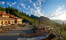 A cozy mountain cabin with a wooden terrace and outdoor seating. In the background, green mountains and a clear sky can be seen.