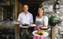 A couple stands in front of a restaurant holding plates with traditional dishes. The atmosphere feels welcoming and rustic.