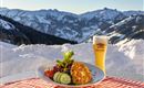 Ein Teller mit einer köstlichen Speise und frischem Gemüse steht auf einem Tisch. Im Hintergrund sind schneebedeckte Berge und ein Bierglas zu sehen.