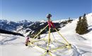 Ein Spielplatz im Schnee mit einem gelben Klettergerüst. Im Hintergrund sind verschneite Berge und ein blauer Himmel zu sehen.