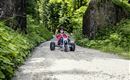 A child is riding a tricycle on a narrow path through a green, wooded environment. The sun is shining, and the mood is cheerful and adventurous.