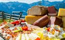 Ein reichhaltiges Käse- und Wurstbrett mit frischen Zutaten und buntem Gemüse. Im Hintergrund sind beeindruckende Berge und ein blauer Himmel zu sehen.