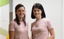 Two women are standing smiling next to each other. Both are wearing pink T-shirts and look friendly and inviting.