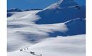 A wintry landscape with snow-covered mountains and a group of ski tourers. The sky is clear and blue, perfect for outdoor activities.