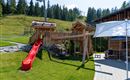 A playground with a red slide, swings, and wooden structures. In the background, green meadows and trees can be seen.