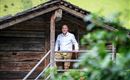 Ein Mann steht auf dem Balkon einer rustikalen Hütte. Im Hintergrund sind grüne Wiesen und Bäume zu sehen.