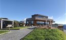 A modern building on a mountain with a clear blue sky and lots of greenery in the background. The area around the building is inviting with outdoor seating options.