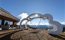 A modern swing set in cloud shape with a view of the mountains. The sky is clear and the sun is shining.