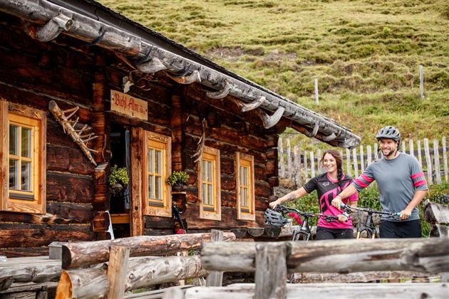 A wooden cabin in a mountainous landscape. Two cyclists stand in front of it, holding their bicycles.