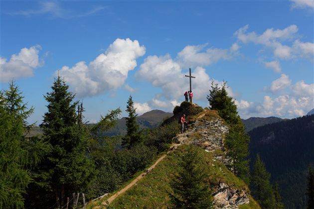A hiker stands next to a summit cross on a mountain ridge, surrounded by green trees. The sky is blue and streaked with white clouds.