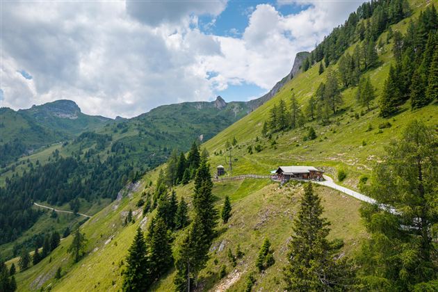 Eine idyllische Berglandschaft mit grünen Wiesen und vereinzelten Bäumen. Im Vordergrund steht eine kleine Hütte, umgeben von Hügeln und Bergen.