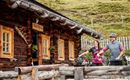 A wooden cabin in a mountainous landscape. Two cyclists stand in front of it, holding their bicycles.