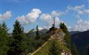A hiker stands next to a summit cross on a mountain ridge, surrounded by green trees. The sky is blue and streaked with white clouds.