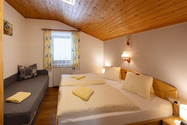 Ein gemütliches Schlafzimmer mit einem Doppelbett, einer Sitzecke und Holzdecke. Die Fenster sind mit bunten Vorhängen geschmückt, und das Licht ist warm und einladend.