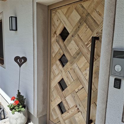 A wooden front door with a decorative pattern. Next to the door stands a flower pot with red flowers and a heart stick.