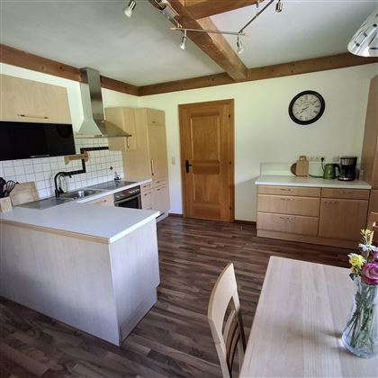 A modern kitchen with wooden shelves and light cabinets. There is a dining table and many windows for natural light.
