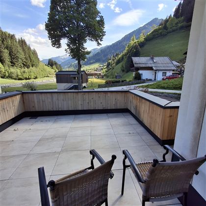 A cozy balcony with two chairs and a view of the mountains and the valley. Surrounded by green forests and a clear sky.