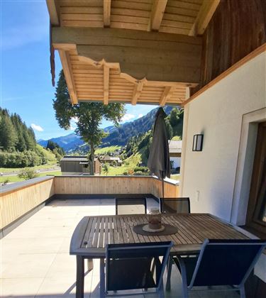 A cozy terrace with a wooden table and chairs. In the background, green mountains and a clear blue sky are visible.