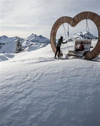 A snowy landscape with mountains in the background. In the foreground, there is a heart-shaped swing with a person sitting on it.
