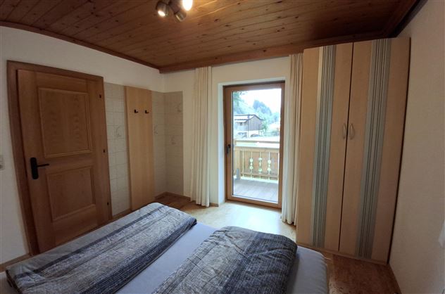 A cozy bedroom with a double bed and wooden ceiling. A window provides access to a balcony with an outdoor view.