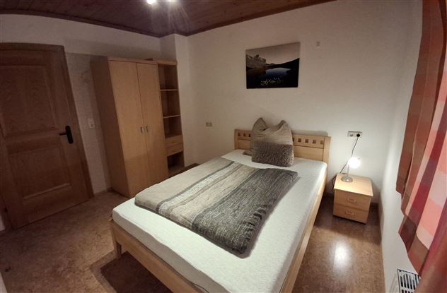 A cozy bedroom with a double bed and a large pillow. There is a wardrobe and a bedside table with a lamp.