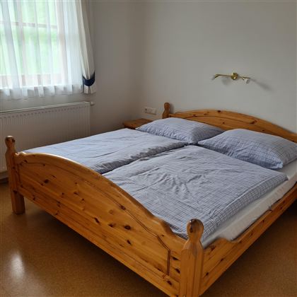 A cozy bedroom with a large wooden bed and gray bedding. The window lets in plenty of light and the walls are simple.