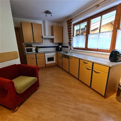 A bright kitchen with wooden cabinets and large windows. A red sofa is positioned in the corner next to the kitchenette.