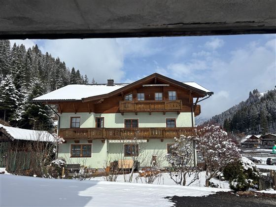 A charming house in Alpine style, surrounded by snow-covered mountains and fir trees. The sky is clear and slightly cloudy.