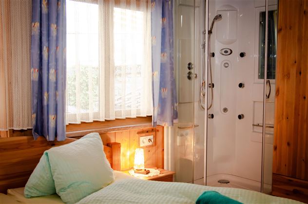 A cozy bedroom with a large window and bright curtains. In the background, there is a modern shower cubicle.