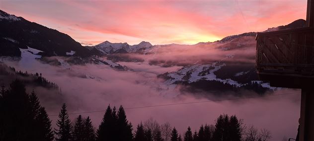 A breathtaking mountain landscape with a colorful sunrise and fog hovering over the valleys. The snow-capped peaks are bathed in warm light.